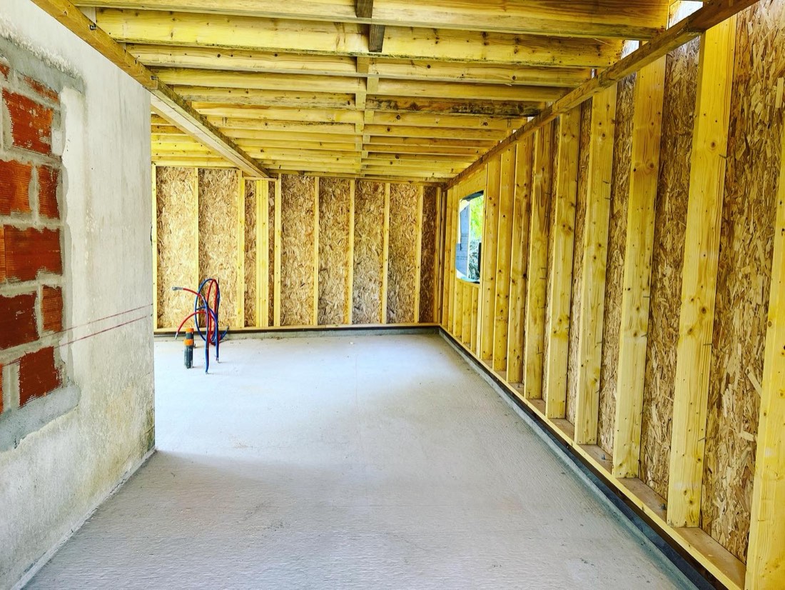 Intérieur d'une extension à ossature bois, montants et panneaux OSB, solivage apparent, raccord visible sur le mur en brique de la maison existante, en Gironde