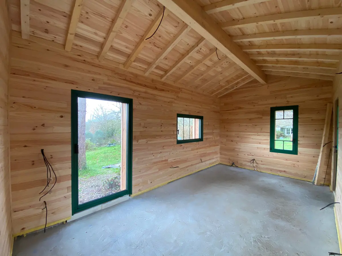 Intérieur d'une construction à ossature bois entièrement habillée en douglas, charpente apparente et porte-fenêtre vitrée ouvrant sur le jardin, chantier de L'Esprit Bois en Gironde