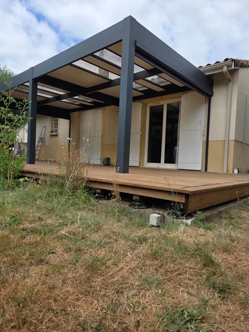 Pergola aluminium anthracite à couverture polycarbonate adossée à une maison, terrasse bois sur plots visible en contrebas, réalisation de L'Esprit Bois à Saint-Pey-de-Castets