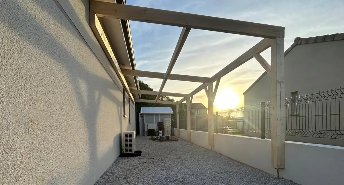 Charpente de carport bois adossée à une façade, vue en contre-jour au coucher du soleil