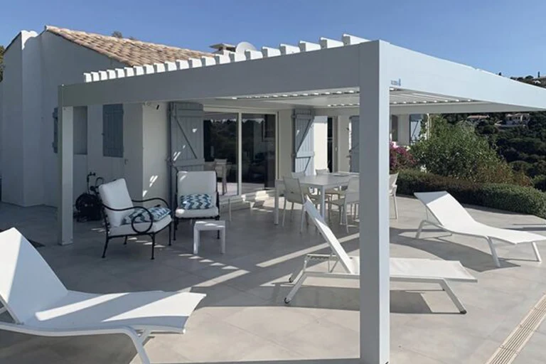 Pergola bioclimatique aluminium blanche adossée à une maison à façade claire, lames orientables
