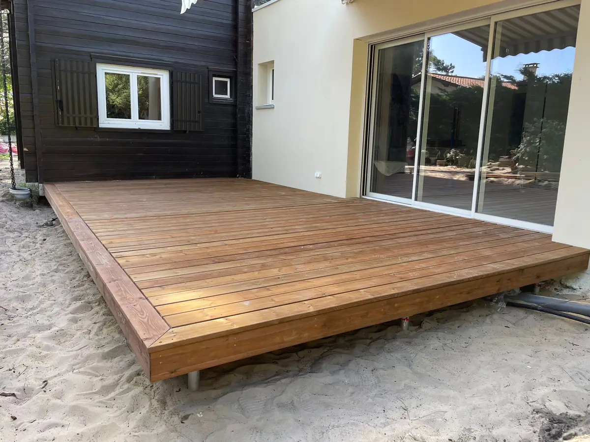 Terrasse en bois sur plots en plein soleil le long d'une maison à bardage noir, réalisation en Gironde