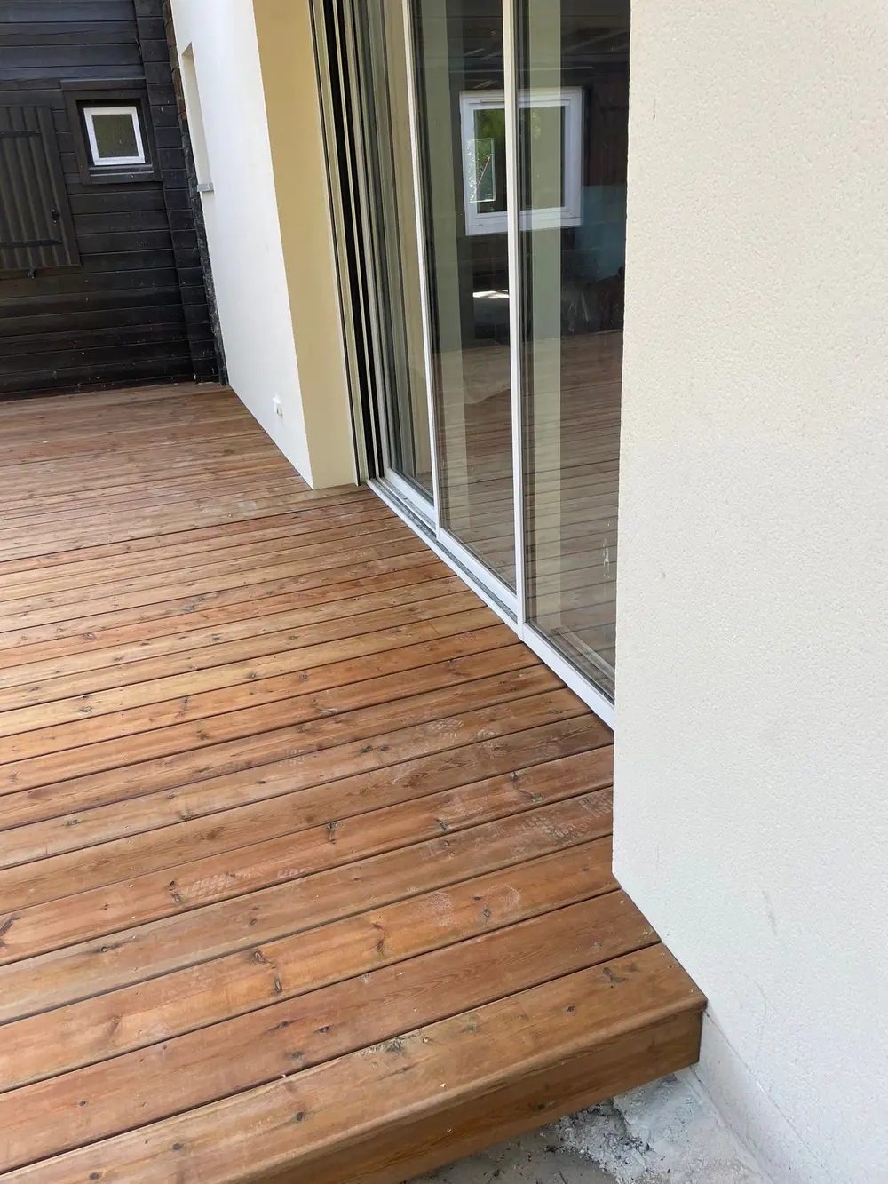 Gros plan sur les lames d'une terrasse en bois au ras du sol, jusqu'à une baie vitrée, avec une marche