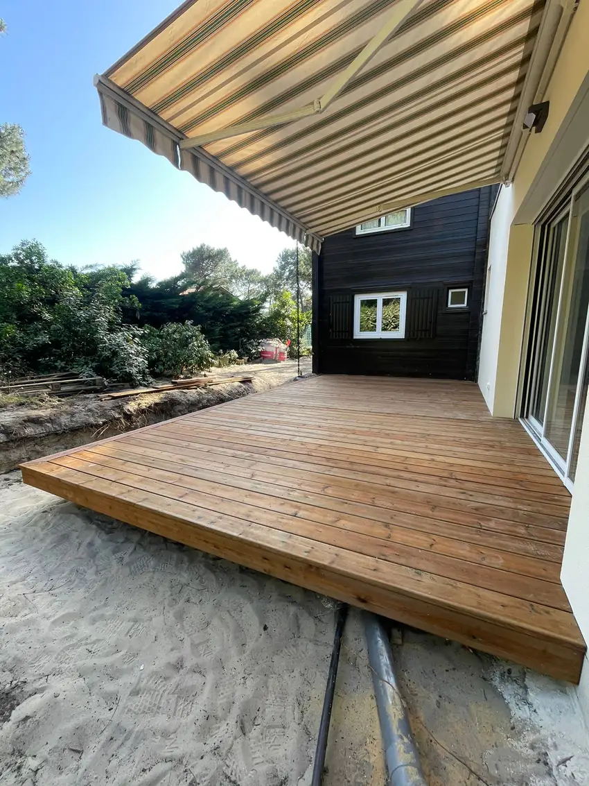 Terrasse en bois protégée par un store banne rayé déployé sous un ciel bleu, en Gironde